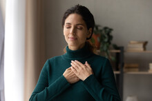 Head Shot Peaceful Young Attractive Woman Holding Folded Hands On Chest, Praying Waiting For Miracle, Feeling Thankful Indoors. Sincere Happy Female Volunteer Showing Kindness Or Expressing Gratitude.
