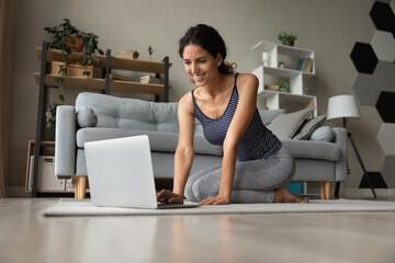 Full length happy millennial generation hispanic latin caucasian woman in active wear sitting on floor carpet with computer, web surfing online training programs, searching internet yoga classes.