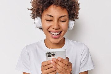 Wall Mural - Cheerful teenage girl smiles hapily uses mobile phone watches funny video smiles broadly uses wireless headphones wears casual t shirt isolated over white background. People entertainment technology