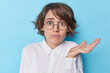 © WHstudio Leushin N - Puzzled hesitant young brunette European woman raises palm with clueless expression cannot make decision wears round spectacles and white formal shirt isolated over blue background. So what to do