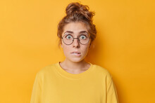 Embarrassed Nervous Woman With Anxious Expression Bites Lips Stars Through Round Spectacles Finds Out Something Dressed In Casual Jumper Isolated Over Yellow Background. Worried Female Model