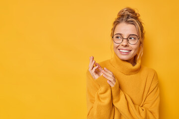 Wall Mural - Horizontal shot of happy young woman rubs hands looks curious awayy smiles toothily wears casual jumper and round spectacles isolated over yellow background blank space for your advertising content