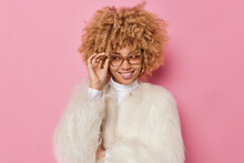 Indoor Shot Of Happy Woman With Bushy Curly Hair Holds Hand On Rim Of Spectacles Smiles Gladfully Looks Away Wears White Winter Coat Isolated On Pink Background. Fashionable Female Model Feels Pleased