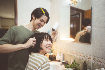 Wall Mural - Young adult asian couple man and woman dressing hair with hairdryer blowing. Together care at home in bathroom.