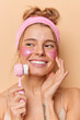© WHstudio Leushin N - Vertical shot of happy woman touches cheek gently smiles broadly uses face massager concentrated away wears pink headband enjoys softness of skin applies patches poses indoor. Spa and beauty