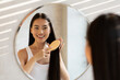 © Prostock-studio - Cheerful asian lady brushing hair, standing in front of mirror