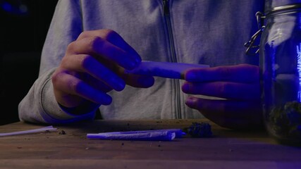 Wall Mural - Caucasian male hands making a cigarette with marijuana. Red and blue police light illuminated criminal hands and glass jar with CBD buds on the table.