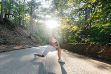 A Young Beautiful Girl Makes Lunges And Warm-up Before Running Training, On The Road In A Dense Forest, During Sunset. Healthy Lifestyle And Running In The Fresh Air. Outdoor Training.