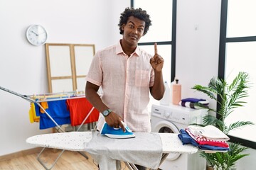 Sticker - African man with curly hair ironing clothes at home pointing finger up with successful idea. exited and happy. number one.