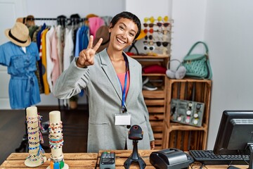 Sticker - Young hispanic woman with short hair working as manager at retail boutique smiling looking to the camera showing fingers doing victory sign. number two.