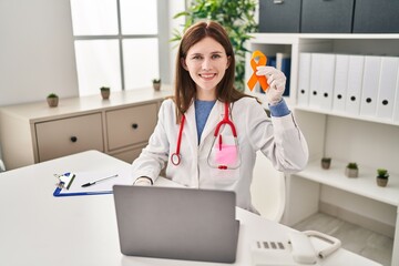 Sticker - Young doctor woman holding awareness orange ribbon looking positive and happy standing and smiling with a confident smile showing teeth