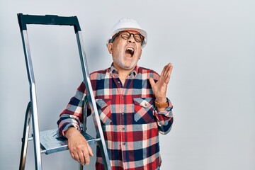 Canvas Print - Handsome mature handyman close to construction stairs wearing hardhat crazy and mad shouting and yelling with aggressive expression and arms raised. frustration concept.