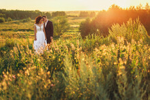 A Couple Of Lovers, A Bride And Groom In A Suit And A Wedding Dress Are Standing In A Field In The Sunset Light, The Groom Is Fooling Around, Joking