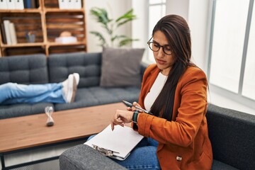 Wall Mural - Young hispanic woman working as psychology counselor checking the time on wrist watch, relaxed and confident