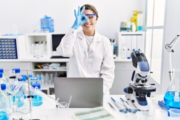 Wall Mural - Young hispanic woman wearing scientist uniform working at laboratory smiling happy doing ok sign with hand on eye looking through fingers