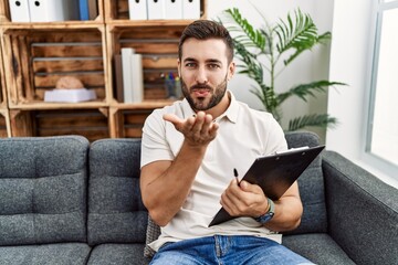 Poster - Handsome hispanic man holding clipboard working at psychology clinic looking at the camera blowing a kiss with hand on air being lovely and sexy. love expression.