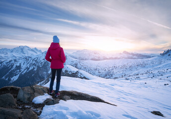 Wall Mural - young woman in red jacket in snowy mountains at sunset in winter. landscape with beautiful girl on t