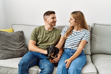 Wall Mural - Young hispanic couple smiling happy sitting on the sofa with dog at home.