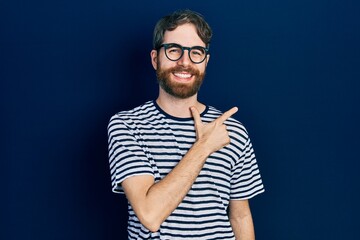 Wall Mural - Caucasian man with beard wearing striped t shirt and glasses cheerful with a smile of face pointing with hand and finger up to the side with happy and natural expression on face