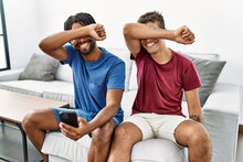 Young Hispanic Men Using Smartphone Sitting On The Sofa At Home Smiling Cheerful Playing Peek A Boo With Hands Showing Face. Surprised And Exited