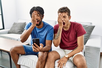 Wall Mural - Young hispanic men using smartphone sitting on the sofa at home yawning tired covering half face, eye and mouth with hand. face hurts in pain.