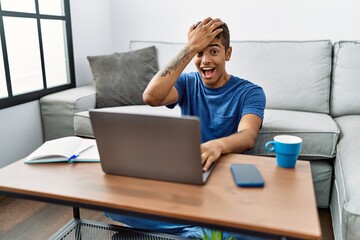 Wall Mural - Young handsome hispanic man using laptop sitting on the floor surprised with hand on head for mistake, remember error. forgot, bad memory concept.