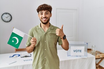 Sticker - Young arab man at political campaign election holding pakistan flag smiling happy and positive, thumb up doing excellent and approval sign