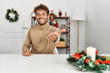 Poster - Young handsome man with beard sitting on the table by christmas decoration smiling cheerful offering palm hand giving assistance and acceptance.