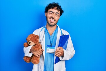 Poster - Young hispanic man wearing doctor uniform holding teddy bear and clipboard sticking tongue out happy with funny expression.