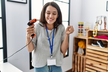 Poster - Young brunette woman working at retail store holding barcode reader device screaming proud, celebrating victory and success very excited with raised arm