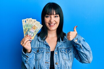 Wall Mural - Young hispanic woman holding 50 romanian leu banknotes smiling happy and positive, thumb up doing excellent and approval sign