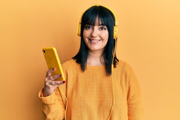 Wall Mural - Young hispanic woman using smartphone wearing headphones looking positive and happy standing and smiling with a confident smile showing teeth