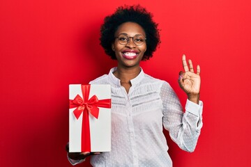 Canvas Print - Young african american woman holding gift doing ok sign with fingers, smiling friendly gesturing excellent symbol