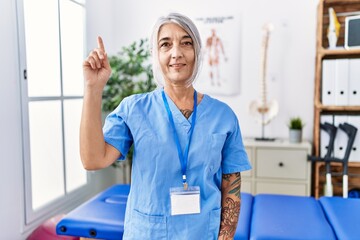 Poster - Middle age grey-haired woman wearing physiotherapist uniform at medical clinic showing and pointing up with finger number one while smiling confident and happy.