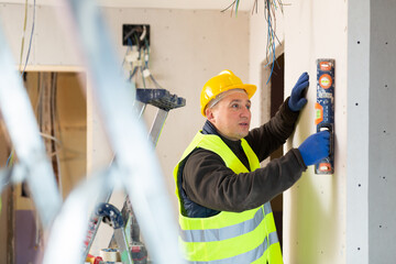 Wall Mural - Repair man in yellow vest and helmet with level in hands measuring evenness of wall.