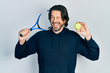 Poster - Middle age caucasian man playing tennis holding racket and ball winking looking at the camera with sexy expression, cheerful and happy face.