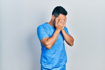 Wall Mural - Handsome hispanic man with beard wearing blue male nurse uniform with sad expression covering face with hands while crying. depression concept.