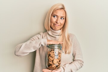 Poster - Young blonde woman holding jar with chocolate chips cookies smiling looking to the side and staring away thinking.