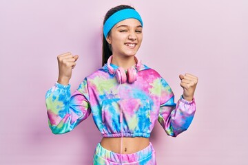 Sticker - Young brunette girl wearing gym clothes and headphones screaming proud, celebrating victory and success very excited with raised arms