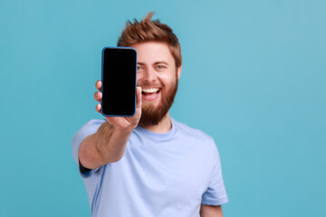 portrait of bearded man holding out mobile phone and looking at camera with toothy smile and positiv
