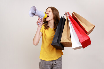 Poster - Young beautiful woman with brown wavy hair wears yellow casual style T-shirt holding shopping bags and megaphone, announcing sale and discount. Indoor studio shot isolated on gray background.