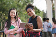 © chachamp - Happy girl student holding tablet and book with friend and looking camera at school park