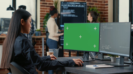 Asian programer writing code in front of computer with green screen chroma key mockup in software development agency. Coder computing big data with team colleagues working online cloud computing.