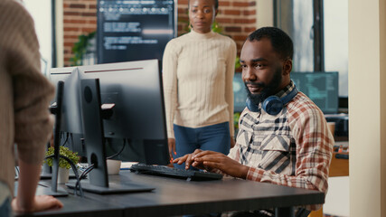 Relaxed software developer wearing wireless headphones smiling working looking at computer screen while typing. Coder writing code in it agency office with colleague asking for informations.
