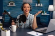 © DC Studio - Portrait of woman smiling at camera in home recording studio. Vlogger with headphones creating content for online social media. Influencer streaming podcast using microphone and laptop.