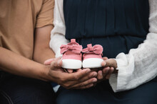 Close Up, Pregnant Women Sit On Bed With Handsome Husband And Hold Couple Red Baby Shoes On The Belly. A Gift For The Baby To Be Born In The Future.