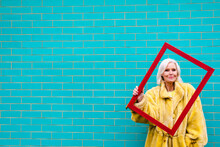 Senior Woman Holding Red Picture Frame Standing In Front Of Turquoise Brick Wall