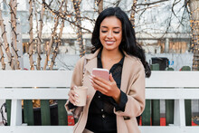 Smiling Woman With Coffee Cup Using Smart Phone On Bench