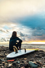Surfer Sitting By Surfboard On Pebbles Looking At Sunset Over Sea