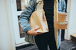 © polinaloves - Young woman wearing protective face mask holding takeaway paper bag with food and bowls near restaurant window. Social distancing during quarantine caused pandemic.
